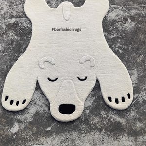 May include: A white polar bear rug with a textured surface. The rug features a bear shape with black accents for the eyes, nose, and paw pads. The rug measures approximately 50 inches in length.