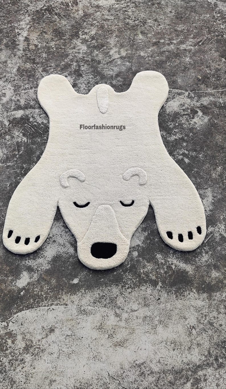 May include: A white polar bear rug with a textured surface. The rug features a bear shape with black accents for the eyes, nose, and paw pads. The rug measures approximately 50 inches in length.