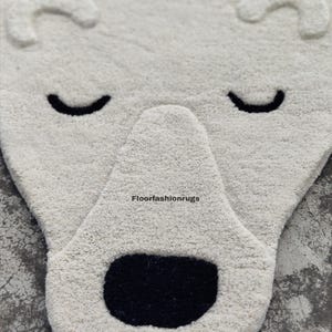 May include: A white, plush rug shaped like a polar bear's face. The rug features a black nose and closed eyes, with curved ears at the top. The rug is designed for a child's room or nursery.