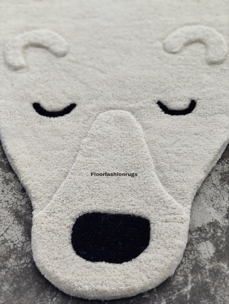May include: A white, plush rug shaped like a polar bear's face. The rug features a black nose and closed eyes, with curved ears at the top. The rug is designed for a child's room or nursery.