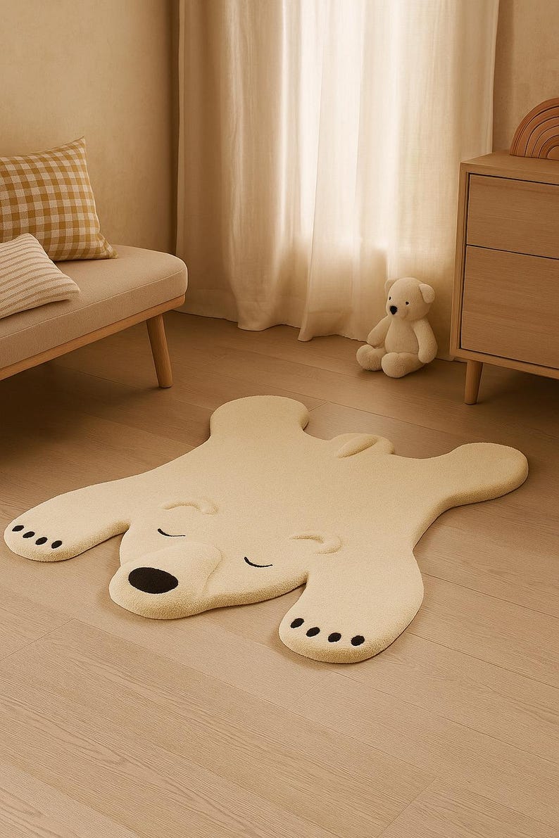 May include: A cream-colored rug shaped like a polar bear, with a black nose and paw pads. The rug is on a light wood floor. A small teddy bear and a beige bench with pillows are in the background.