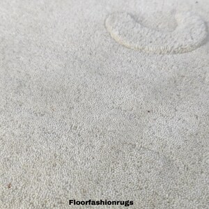 May include: A cream-colored rug with a textured surface and a bear-shaped design. The rug features a bear face with black eyes and a rounded ear. The text "Floorfashionrugs" is at the bottom.
