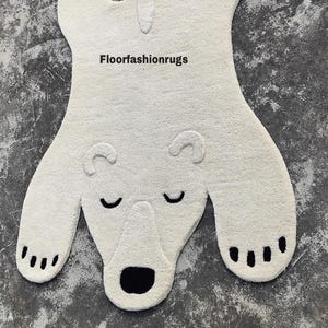 May include: A cream-colored polar bear rug with a black nose, closed eyes, and paw pads. The rug has a soft, textured appearance and is shaped like a bear. The text "Floorfashionrugs" is visible on the rug.