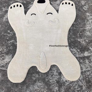 May include: A cream-colored rug shaped like a polar bear, with black accents for the eyes, nose, and paw pads. The rug has a soft, plush texture and is designed with a playful, cartoonish aesthetic. The rug is a decorative floor covering.