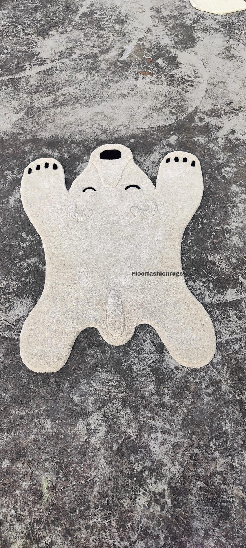 May include: A cream-colored rug shaped like a polar bear, with black accents for the eyes, nose, and paw pads. The rug has a soft, plush texture and is designed with a playful, cartoonish aesthetic. The rug is a decorative floor covering.