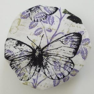 May include: A round fabric cover featuring a white background with a black butterfly print and purple botanical illustrations. The textile has a textured weave and displays a repeating pattern of butterflies and leaves across the circular surface.