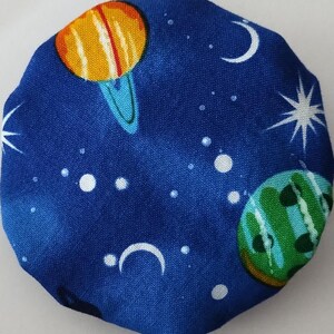 May include: A blue fabric item with a celestial design. The pattern includes planets in shades of orange, green, and blue, along with white stars, moons, and small white dots. The item has an octagonal shape.