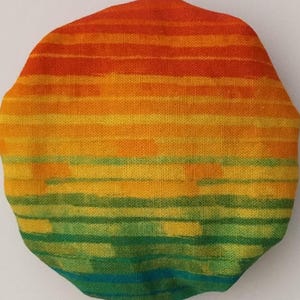 May include: A round, colorful fabric with a striped pattern in shades of red, orange, yellow, green, and blue. The fabric has a textured surface and appears to be made of a woven material.