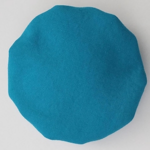 May include: A circular, teal-colored fabric cap with a gathered, elasticized edge. The material has a visible woven texture and a solid, uniform color throughout the entire surface of the rounded, dome-shaped head covering accessory.