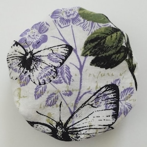 May include: A round fabric-covered button featuring a white background with a printed pattern of two black-outlined butterflies, purple flowers, green leaves, and faint cursive script text scattered across the surface of the textile material.