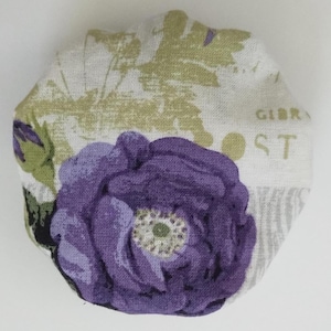 May include: A circular fabric-covered button featuring a prominent purple flower with a white and green center. The background fabric is off-white with muted green botanical leaf patterns and partial text that includes the letters GIBR and POST.