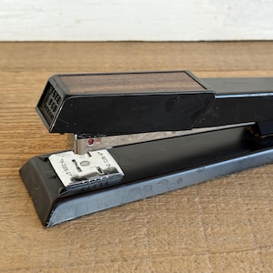May include: Black stapler with a wooden top. The stapler has a rectangular base and a lever for stapling. The stapler is made of metal and has a silver staple guide. The stapler is on a wooden surface.