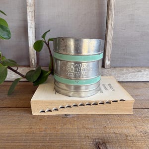 May include: A vintage metal sifter with a mint green band and the text "Savory Gift-Chime". The sifter is on a book. A green plant is in the background. The sifter is a kitchen utensil.
