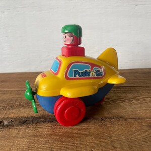 May include: A vintage toy airplane in yellow, blue, and red. The toy has a green propeller, red wheels, and a small figure on top. The plane has the words "Push'n'Go" on the side.