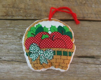 May include: A fabric ornament shaped like a basket filled with red apples with white polka dots. The basket has a brown woven pattern, green holly leaves, and a green and white bow. A red yarn loop is attached for hanging.