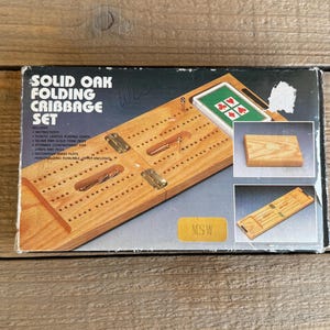 Op de afbeelding: Een massief eikenhouten opvouwbare cribbage set in een doos. De set bevat een houten cribbagebord met messing scharnieren, pinnen en een kaartspel. De doos heeft de tekst "SOLID OAK FOLDING CRIBBAGE SET".