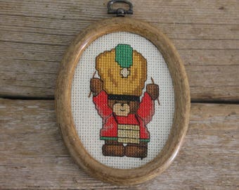 May include: Oval-framed cross-stitch featuring a teddy bear in Native American attire. The bear wears a yellow headdress with a green stripe, a red tunic, and brown boots. The frame is made of light brown wood.