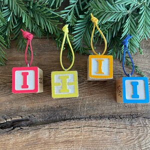 May include: Four wooden block ornaments with the letter "I" painted on each. The blocks are painted in red, yellow, blue, and natural wood. Each block has a string loop for hanging.