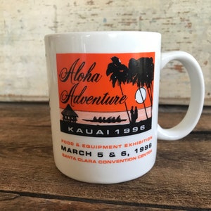 May include: White ceramic mug with an orange and black design. The design features palm trees, a sunset, and the text "Aloha Adventure Kauai 1996". The text "Food & Equipment Exhibition March 5 & 6, 1996 Santa Clara Convention Center" is also included.