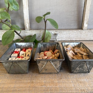 May include: Three vintage metal containers filled with game pieces. One holds red and white letter tiles, another has red and tan heart-shaped pieces, and the third contains wooden letter tiles. The containers sit on a wooden surface.