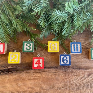 May include: Nine wooden block ornaments with painted numbers and symbols. The ornaments are in various colors, including red, yellow, blue, and green. The blocks are hanging from a string.