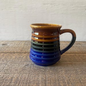 May include: A handmade ceramic mug with a ribbed design and a curved handle. The mug features a gradient of colors, including brown, orange, green, black, and blue. The mug is sitting on a wooden surface.
