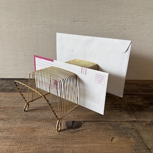 May include: A gold-colored metal letter holder with a wire design. The holder is filled with white envelopes and a pink and white card. The holder sits on a wooden surface.