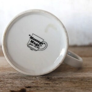 K&ouml;nnte beinhalten: Wei&szlig;e Keramik-Kaffeetasse mit einem schwarzen Logo, das "Message Mugs" lautet, und einem kleinen Logo, das "Joy 1993" lautet.