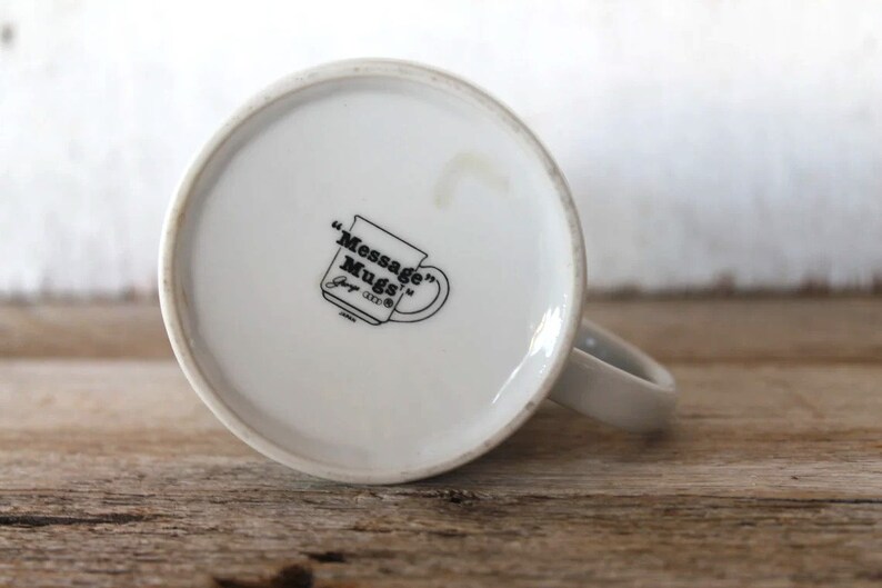 K&ouml;nnte beinhalten: Wei&szlig;e Keramik-Kaffeetasse mit einem schwarzen Logo, das "Message Mugs" lautet, und einem kleinen Logo, das "Joy 1993" lautet.
