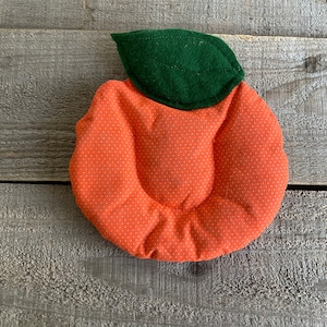 May include: Orange, quilted fabric coaster with white polka dots, shaped like a peach. A green felt leaf is attached to the top. The coaster is placed on a weathered, gray wooden surface. This item is suitable for kitchen and home decor.