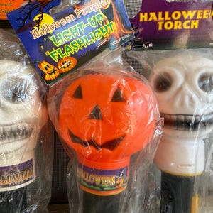May include: Three Halloween-themed light-up flashlights. One is a white skull, another is an orange pumpkin, and the third is a white skull. Each is packaged in clear plastic. The pumpkin flashlight has the text "Halloween Pumpkin LIGHT-UP FLASHLIGHT".