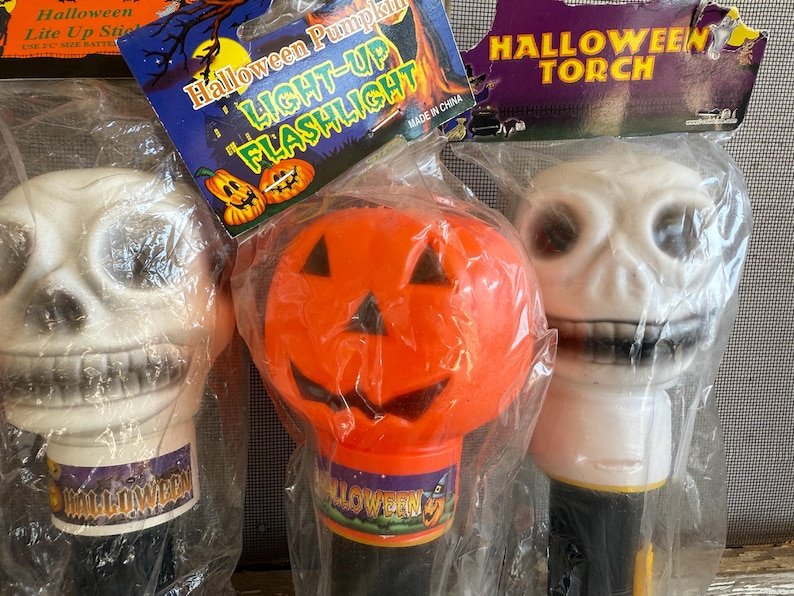 May include: Three Halloween-themed light-up flashlights. One is a white skull, another is an orange pumpkin, and the third is a white skull. Each is packaged in clear plastic. The pumpkin flashlight has the text "Halloween Pumpkin LIGHT-UP FLASHLIGHT".
