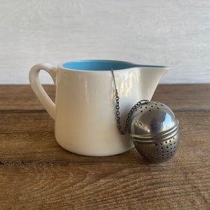 May include: A cream-coloured creamer with a light blue interior and a silver tea infuser. The creamer has a handle and a spout. The tea infuser is a perforated sphere attached to a chain.