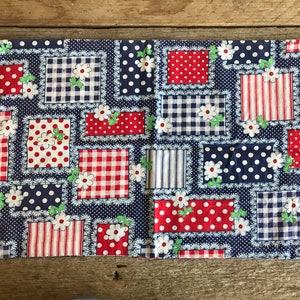 May include: A blue and white patchwork fabric placemat with red and white gingham, polka dot, and striped patterns. The placemat features white flowers with green leaves.
