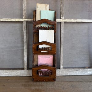 May include: A vintage wooden mail organiser with three compartments, each labelled with carved lettering. The organiser holds various papers and envelopes, including a pink form. The background features a weathered window frame.