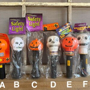 May include: A collection of Halloween-themed flashing safety lights and torches. The lights feature pumpkin and skull designs, with black handles and clear plastic packaging. Some lights have the words "Safety Light" on the packaging.
