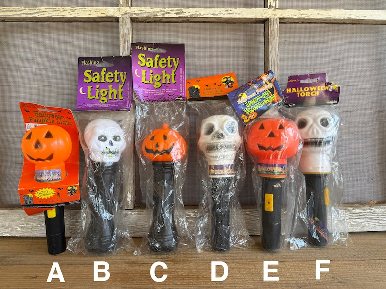 May include: A collection of Halloween-themed flashing safety lights and torches. The lights feature pumpkin and skull designs, with black handles and clear plastic packaging. Some lights have the words "Safety Light" on the packaging.