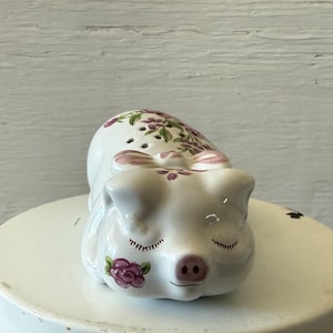 Pode incluir: Estatueta de porco em cerâmica branca com olhos fechados, decorada com detalhes florais rosa e roxos. O porco está deitado, com um pequeno laço nas costas. A estatueta está sobre uma superfície branca.