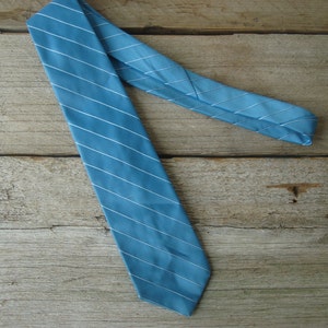 May include: A teal-colored necktie with thin white stripes. The tie is made of a smooth fabric and is diagonally positioned against a wooden background. The tie is a classic accessory for formal or business attire.