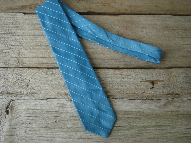 May include: A teal-colored necktie with thin white stripes. The tie is made of a smooth fabric and is diagonally positioned against a wooden background. The tie is a classic accessory for formal or business attire.
