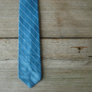 May include: A blue tie with white diagonal stripes against a wooden background. The tie is a solid light blue color with thin white stripes running from the top left to the bottom right. The background is made of wooden planks.