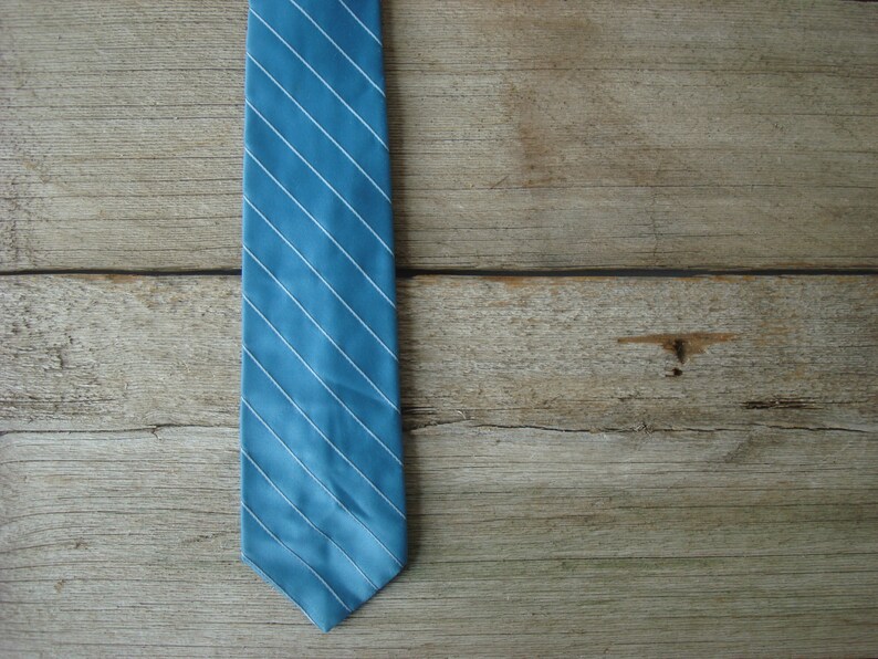May include: A blue tie with white diagonal stripes against a wooden background. The tie is a solid light blue color with thin white stripes running from the top left to the bottom right. The background is made of wooden planks.