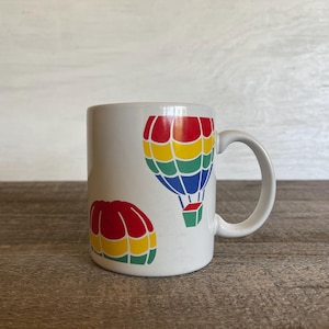 Puede incluir: Taza de cerámica blanca con un diseño de globo aerostático colorido. El globo es rojo, amarillo, azul y verde. Hay un globo aerostático más pequeño en la esquina inferior izquierda de la taza.