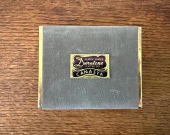 Könnte beinhalten: Eine Vintage-Canasta-Kartenspielbox. Die Box ist grau mit goldenem Rand und einem rechteckigen Etikett mit der Aufschrift "Plastic Coated Duratone Playing Cards Canasta". Die Box liegt auf einer Holzoberfläche.