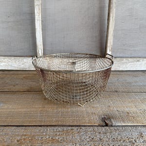 May include: A round, metal wire basket with a handle on each side. The basket is made of a fine mesh and has a slightly reddish-brown hue. It sits on a weathered wooden surface, with a window frame in the background.