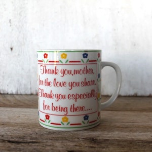 K&ouml;nnte beinhalten: Wei&szlig;e Keramiktasse mit rotem, gr&uuml;nem, blauem und gelbem Blumenrand. Die Tasse hat eine rote Aufschrift mit dem Text: "Danke, Mutter, f&uuml;r die Liebe, die du teilst. Danke besonders daf&uuml;r, dass du da bist..."
