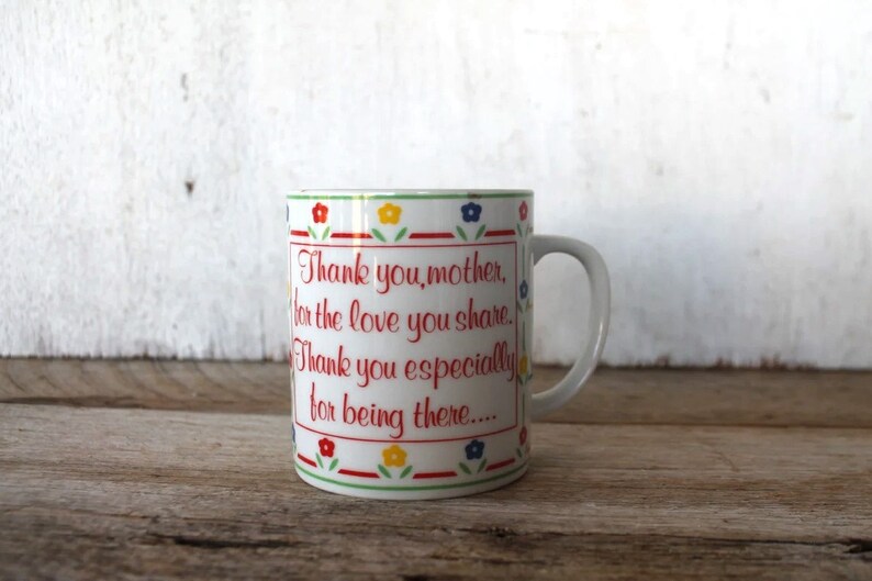 K&ouml;nnte beinhalten: Wei&szlig;e Keramiktasse mit rotem, gr&uuml;nem, blauem und gelbem Blumenrand. Die Tasse hat eine rote Aufschrift mit dem Text: "Danke, Mutter, f&uuml;r die Liebe, die du teilst. Danke besonders daf&uuml;r, dass du da bist..."