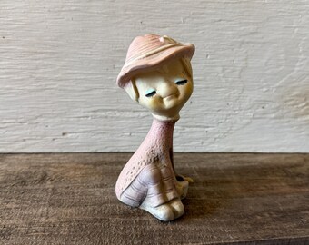 Könnte beinhalten: Vintage Keramikfigur einer sitzenden Figur mit rosa Hut und Kleid. Die Figur hat einen ruhigen Ausdruck mit geschlossenen Augen und einem leichten Lächeln. Die Farbpalette umfasst sanfte Rosatöne, Cremes und einen Hauch von Braun.