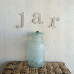 Könnte beinhalten: Ein hellblaues Glasgefäß mit einem Korkstopfen, das auf einer geflochtenen Weidenoberfläche steht. Das Wort "jar" ist in weißen Buchstaben auf einer weißen Wand hinter dem Gefäß geschrieben.