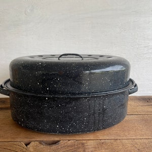 May include: A black speckled enamelware roasting pan with a lid. The lid has a handle and small holes. The pan sits on a wooden surface. The pan is designed for cooking.